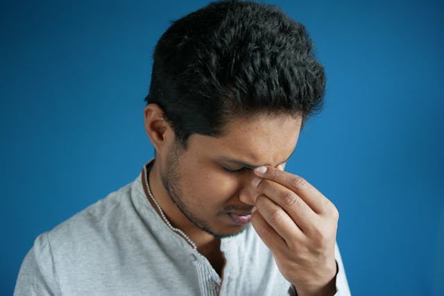 A man showing stress or headache by pinching his nose bridge against a blue backdrop.