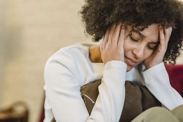 A woman with curly hair sits indoors, appearing stressed and holding her head in her hands.