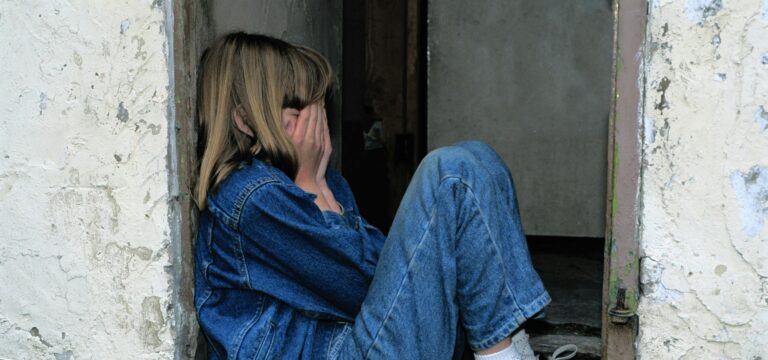 A young girl in denim sits alone in a doorway looking distressed, conveying loneliness.