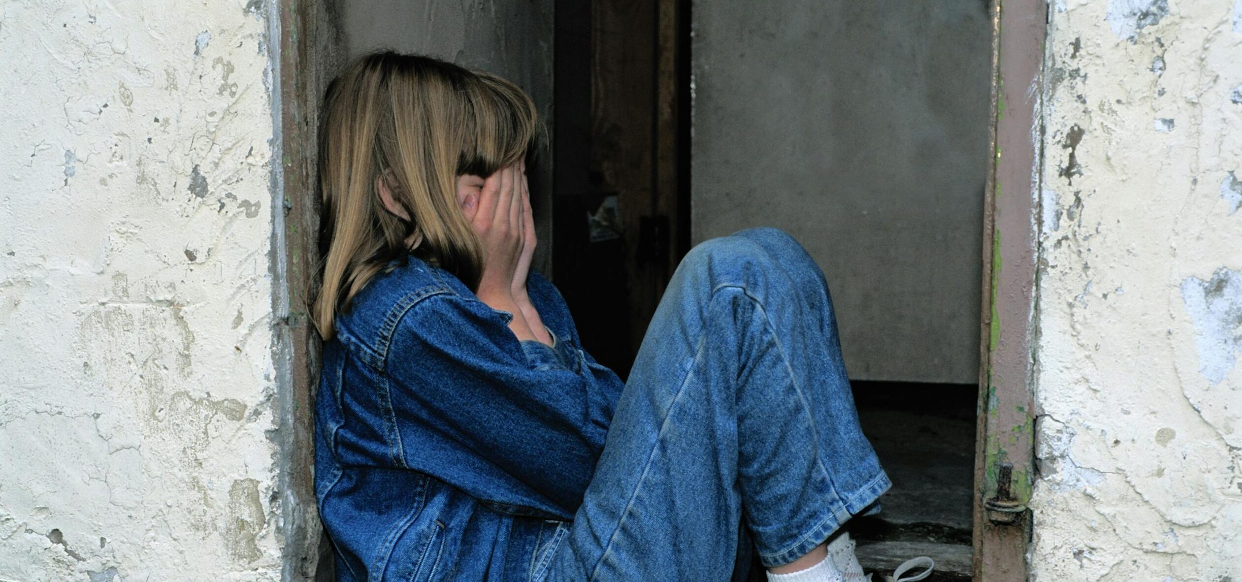 A young girl in denim sits alone in a doorway looking distressed, conveying loneliness.