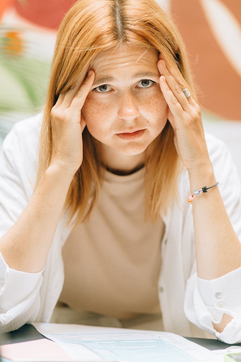 Close-up of a woman sitting at a desk with hands on her head, looking stressed and exhausted.
