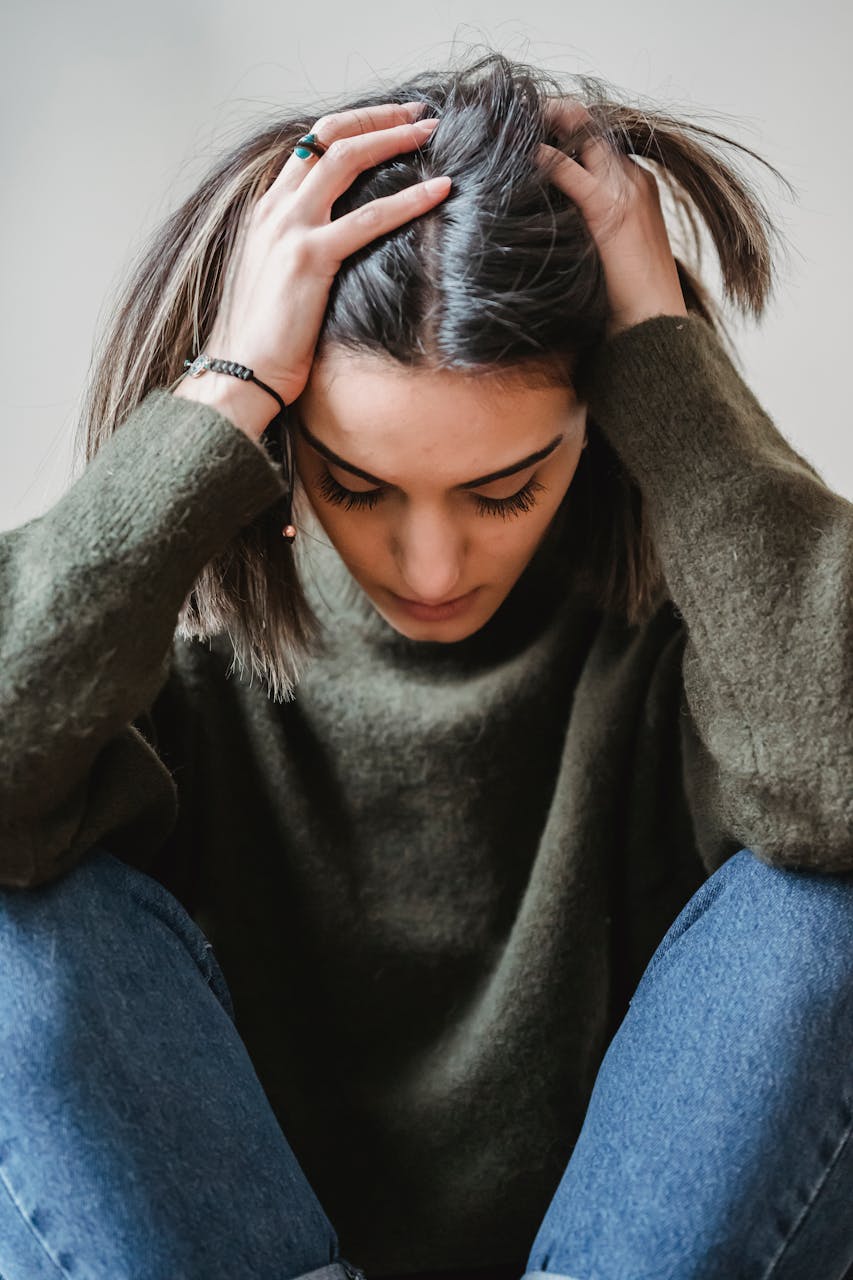 Desperate female in casual clothes touching head while sitting near white wall with bent legs in light room at home