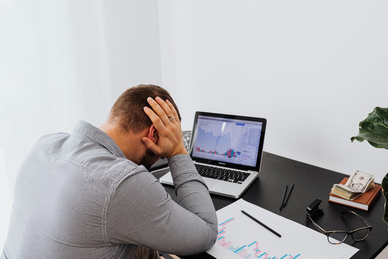 A businessman holds his head in frustration while sitting at a desk with a laptop and financial charts.