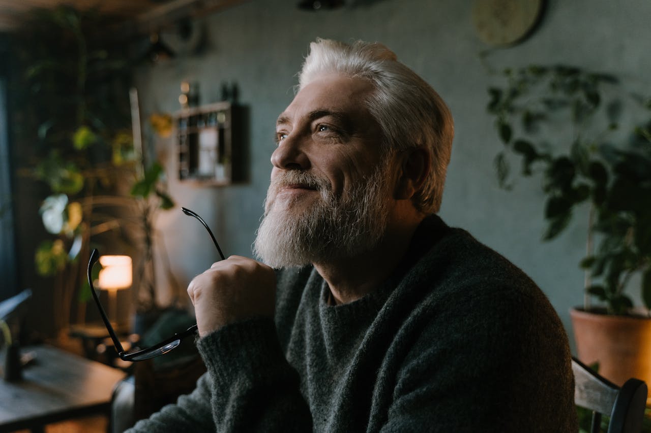A contemplative senior man with gray hair holding eyeglasses, enjoying a serene moment indoors.