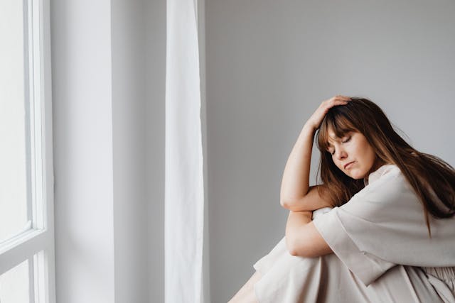 A woman in white sits pensively by a window, reflecting solitude indoors.
