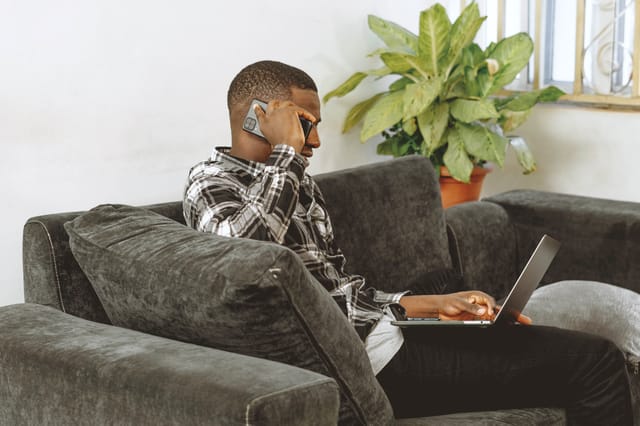 A young man multitasking with a smartphone and laptop on a sofa, illustrating remote work from home.