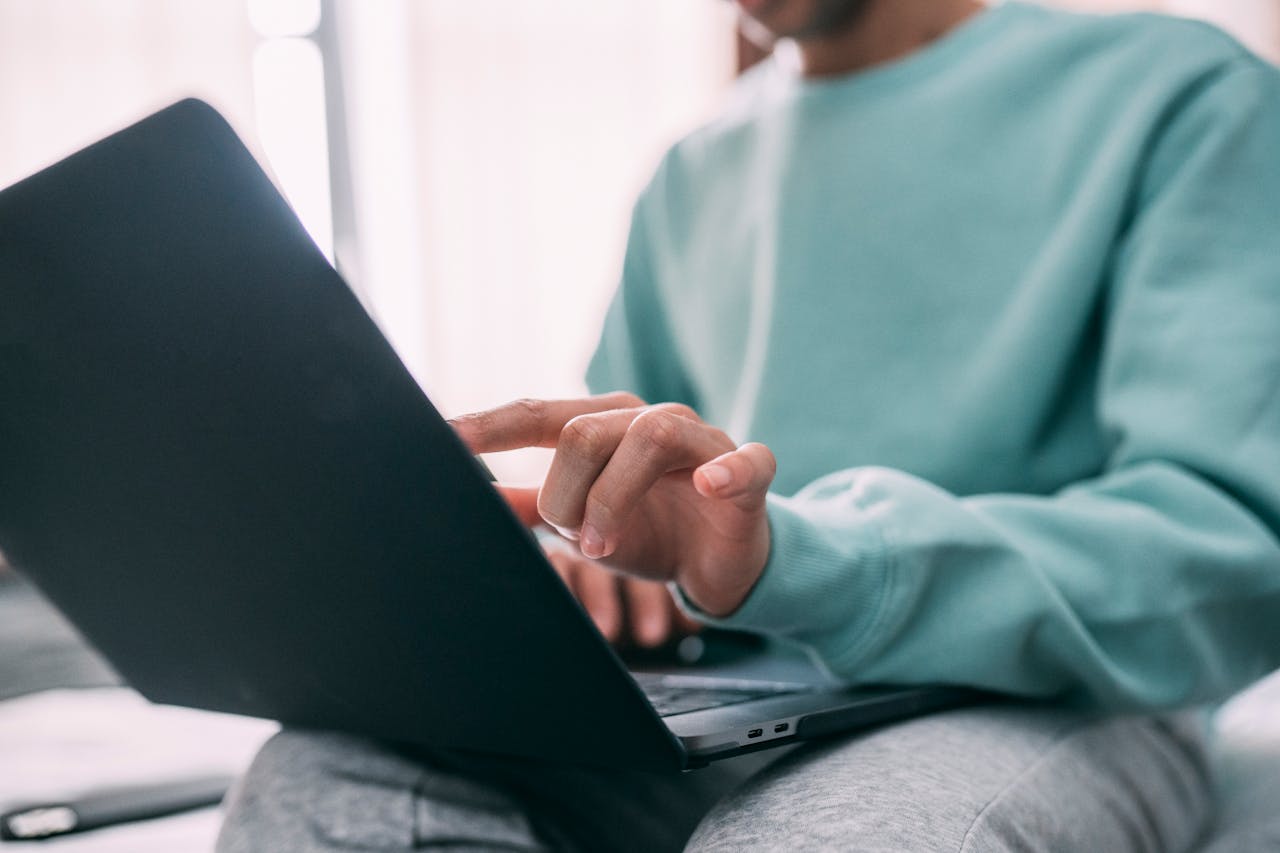 Crop male wearing casual clothes sitting on couch with laptop on knees in light room with large window while touching screen with finger searching for the best OCD clinic
