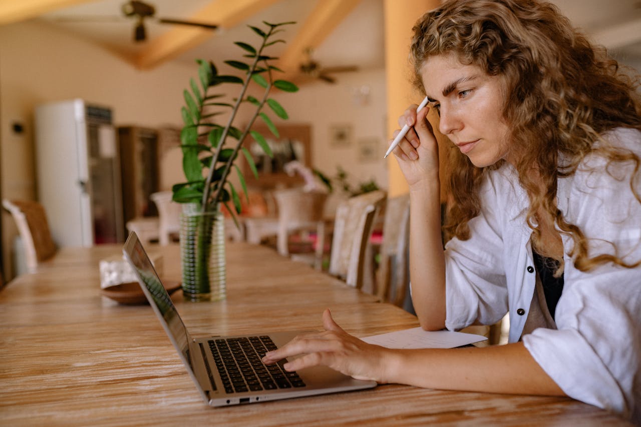 Focused woman with curly hair using laptop at home table, multitasking and working remotely.
