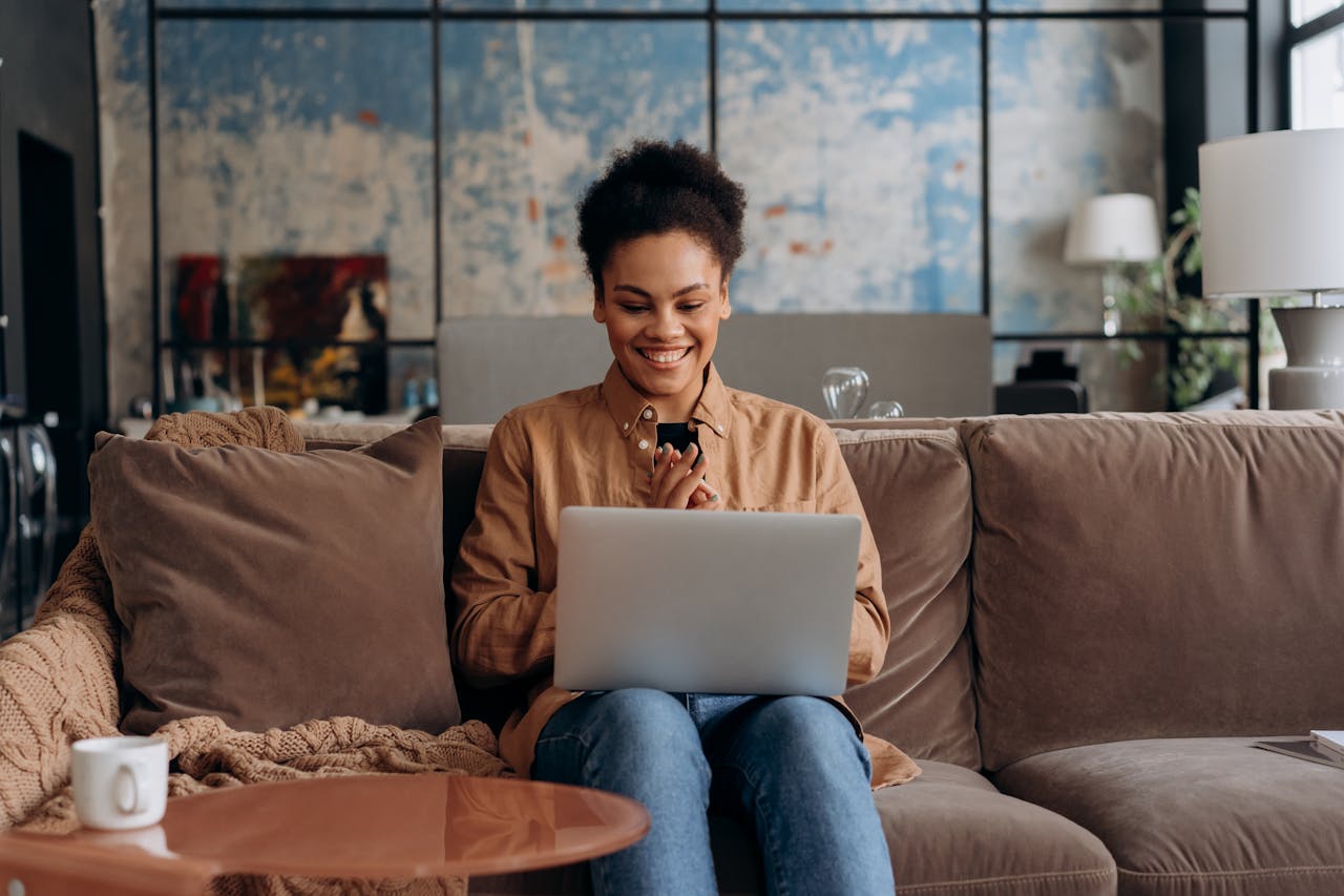 Smiling woman sits on a couch using a laptop, enjoying her home environment.