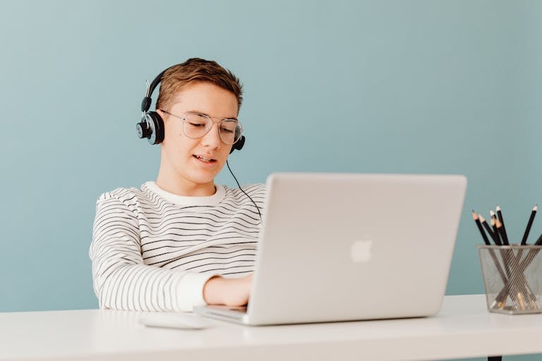 Teenager with glasses using a laptop and headphones, sitting at a table.