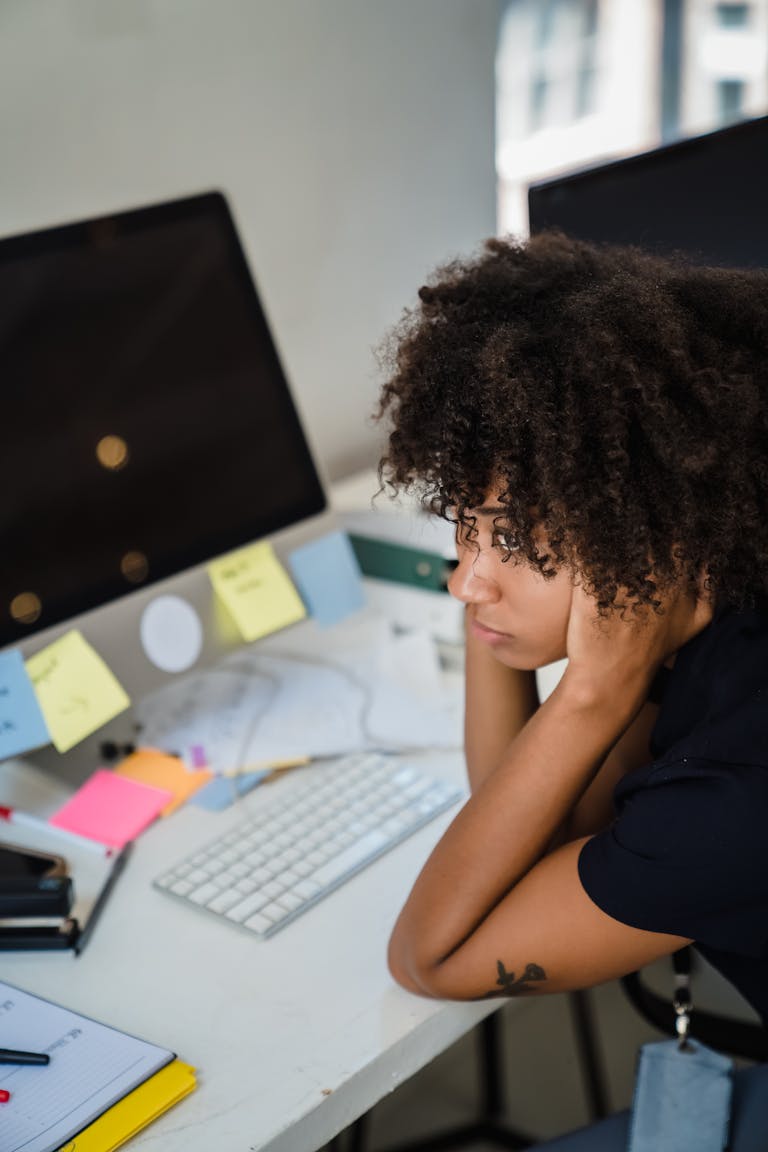 Woman with afro hair appearing stressed while sitting at her home office desk, surrounded by computers and sticky notes.