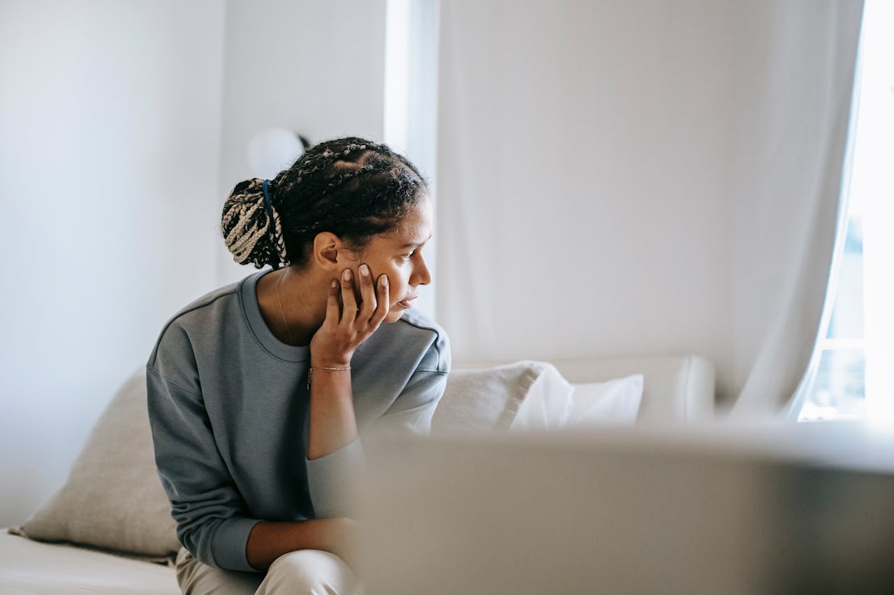Young black female in casual outfit sitting on cozy white bed and thinking about finding an ERP therapist