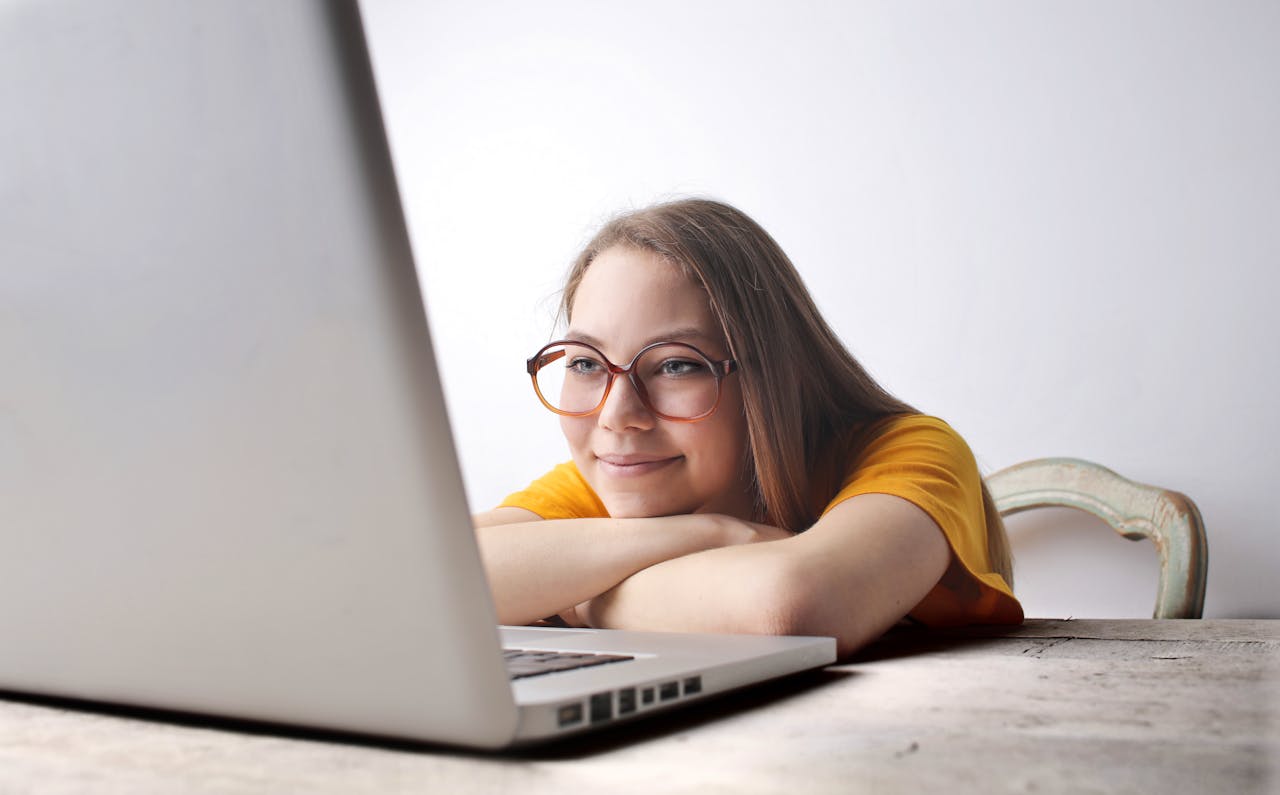 Young woman with glasses smiles while using a laptop at a desk indoors.