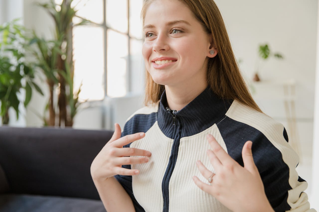 A cheerful young woman sitting indoors, smiling with casual attire in a bright living room.