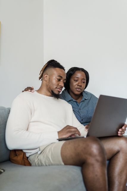 African American couple sitting on a couch using a laptop together, enjoying a cozy moment at home.