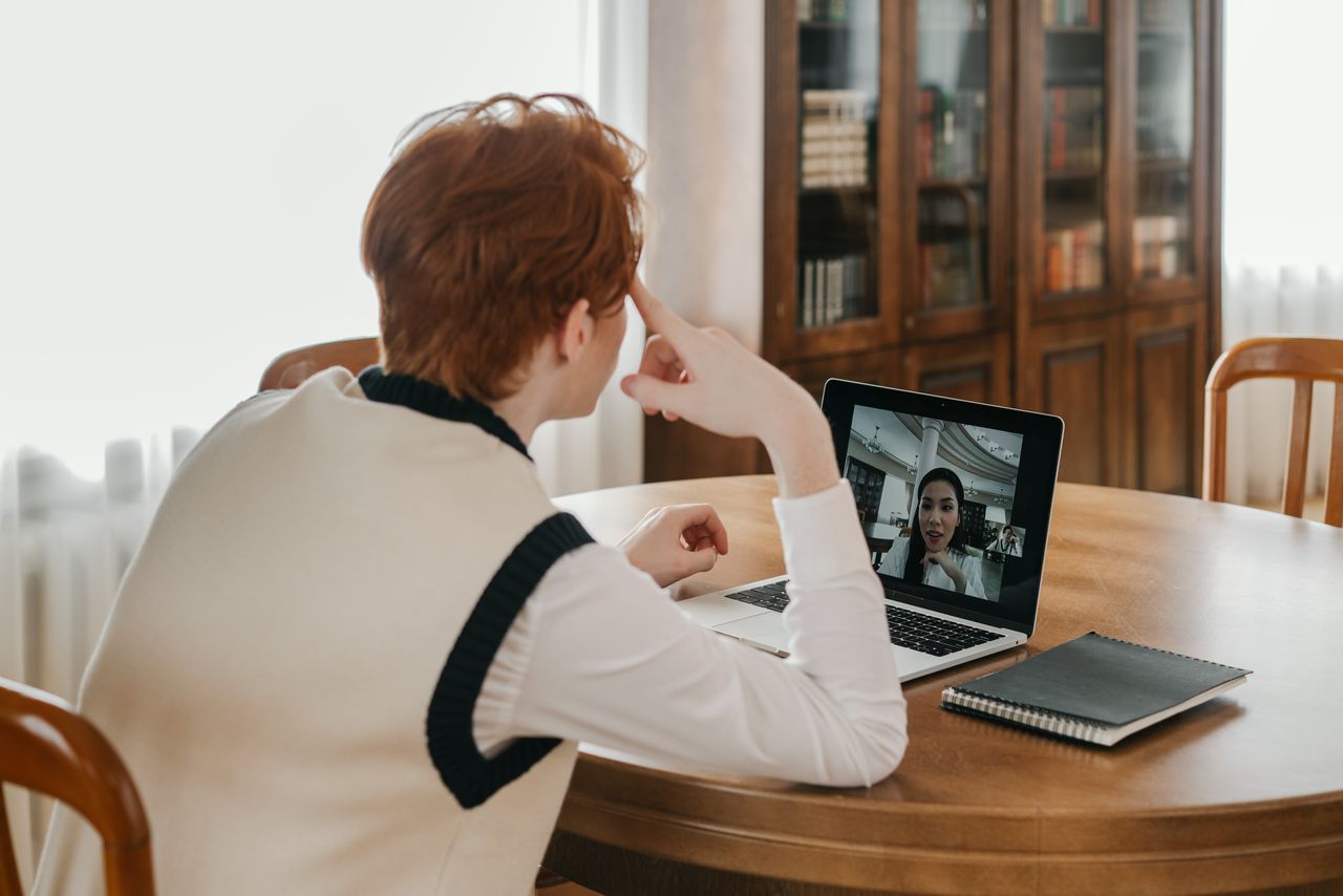 A man sitting at a table, using a laptop for a video call, set in a cozy indoor space.