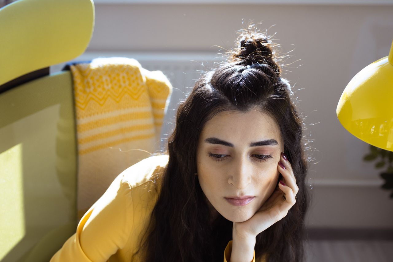 A woman in a yellow setting, hand on chin, looking contemplative indoors.