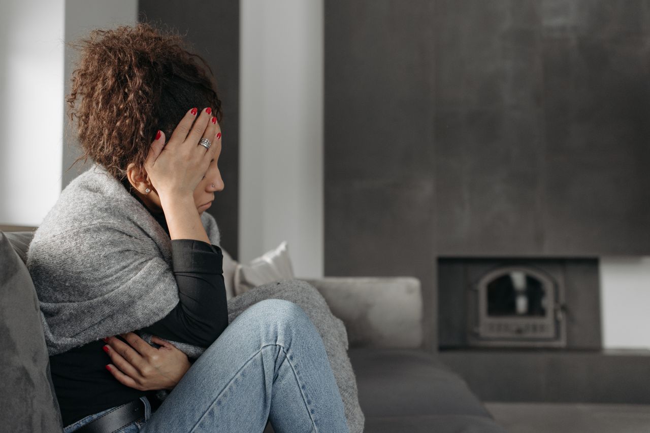 A woman sits on a couch indoors, wrapped in a blanket, holding her head in discomfort.