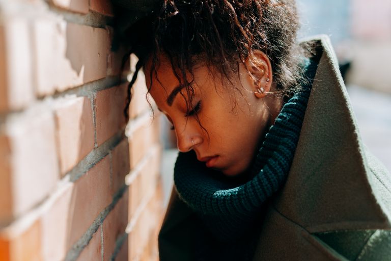 Young woman in a green coat leans against a brick wall, eyes closed, capturing introspection and solitude.