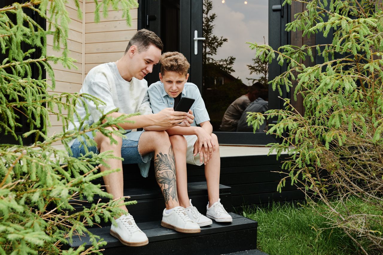 Father and son sitting outdoors sharing a moment on a smartphone.