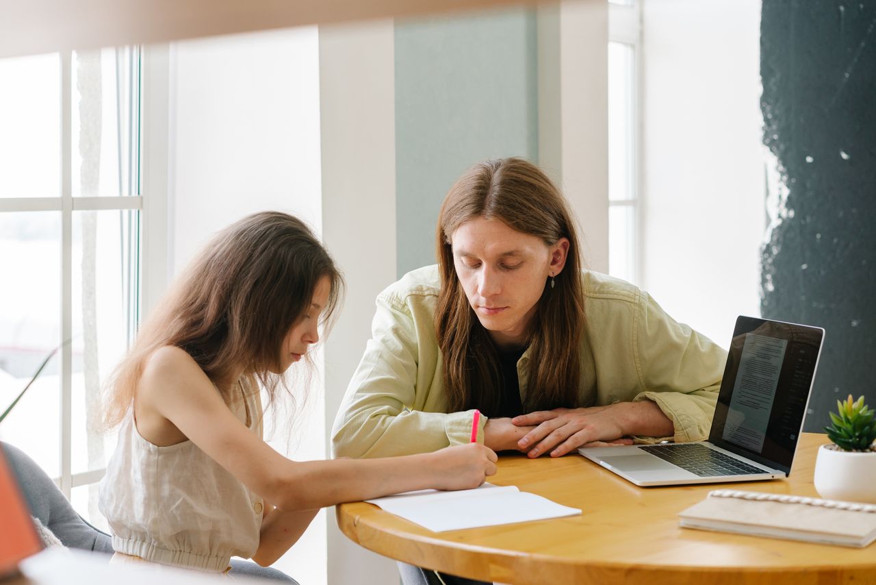 A father helps his daughter with homeschooling indoors, featuring a laptop and study materials.
