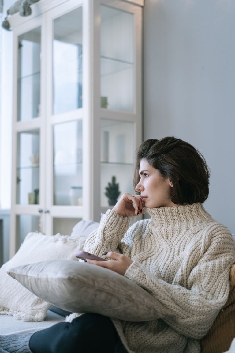A thoughtful woman in a knitted sweater holding a smartphone while sitting indoors on a couch.