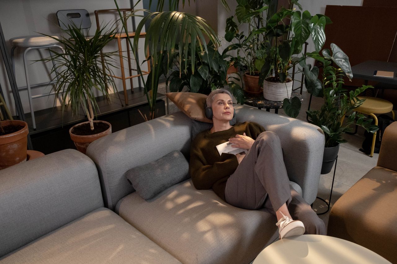Mature woman with short gray hair relaxing on a couch with headphones, surrounded by lush indoor plants.